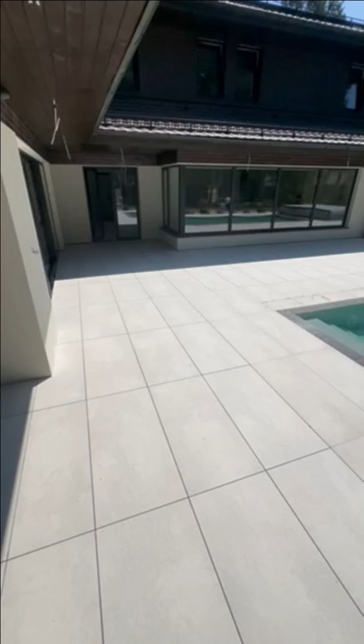 Duży taras z jasnych płyt gresowych przy nowoczesnym domu jednorodzinnym z przeszkloną elewacją i basenem w ogrodzie.