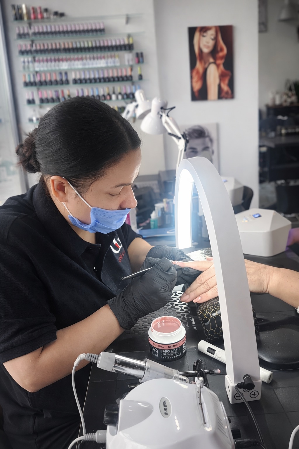 Stylistka paznokci wykonuje manicure hybrydowy klientce pod lampą, precyzyjnie opracowując płytkę paznokcia w salonie kosmetycznym