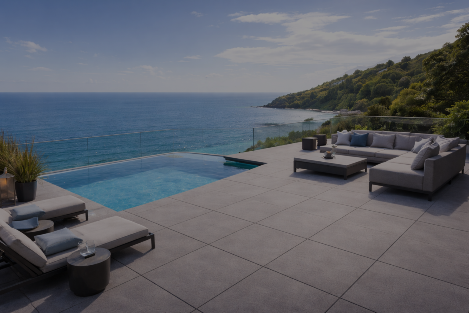 Nowoczesny taras z dużych płyt gresowych przy domu na klifie, z basenem infinity, leżakami i sofą wypoczynkową oraz widokiem na morze i zielone wzgórza.