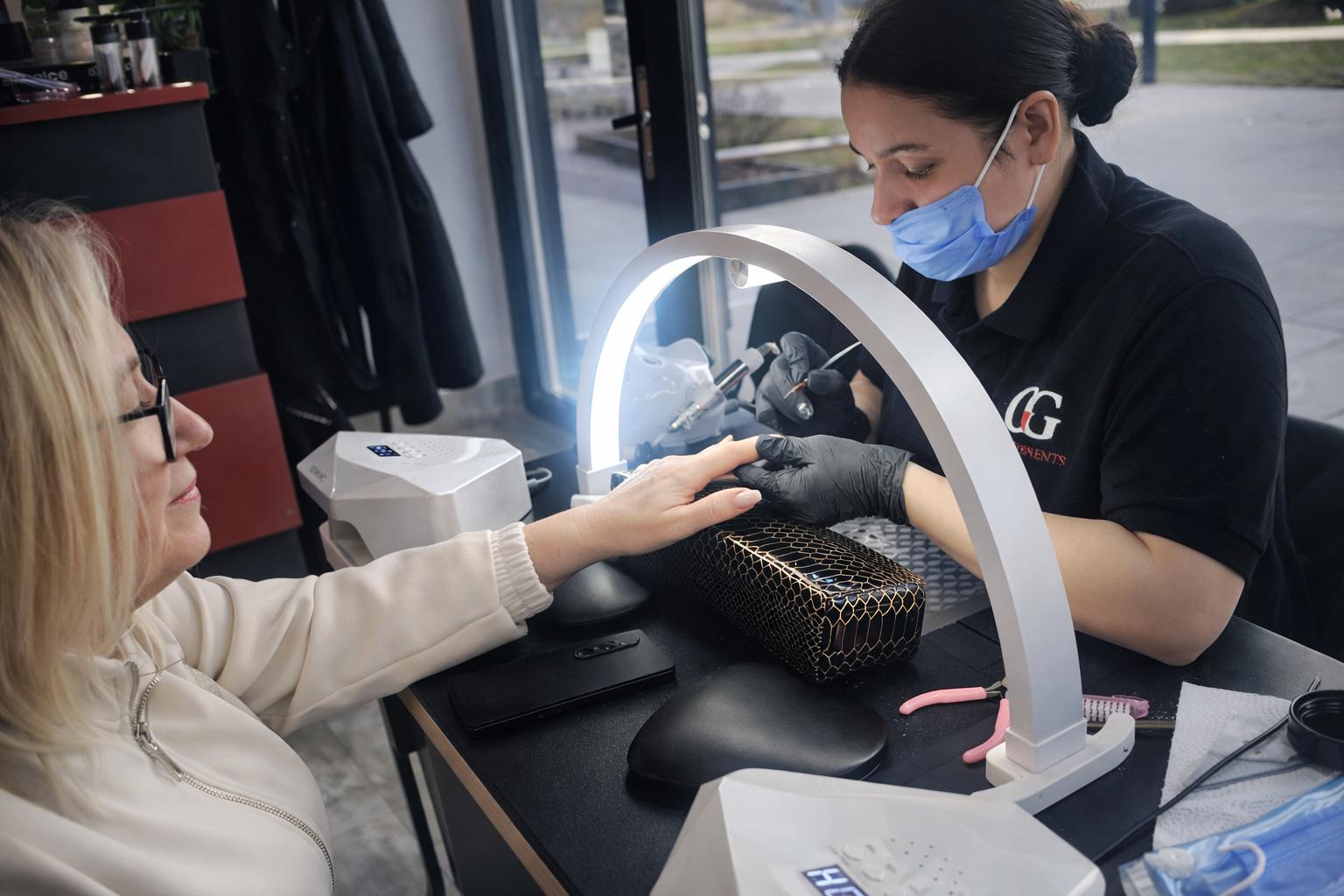 Stylistka wykonuje manicure klientce przy użyciu lampy i narzędzi, pracując w profesjonalnym salonie kosmetycznym