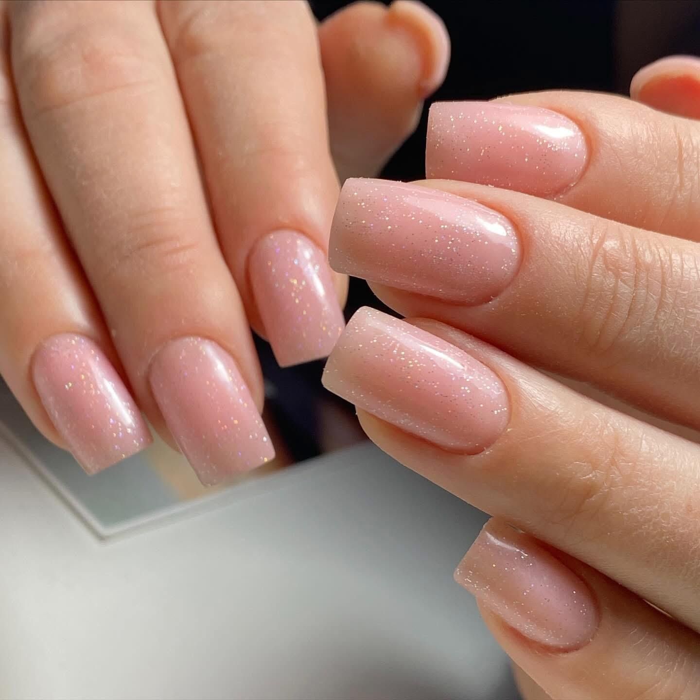 Dłonie z paznokciami w delikatnym różowym odcieniu z subtelnymi drobinkami, tworzące elegancki i naturalny manicure