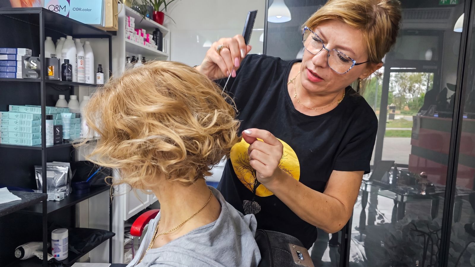 Fryzjerka dopracowująca krótką blond fryzurę z lokami podczas stylizacji w salonie fryzjerskim.