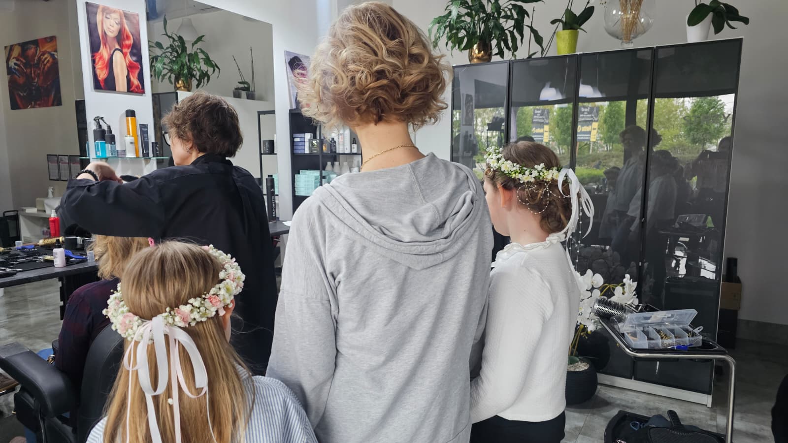 Dziewczynki w komunijnych fryzurach z kwiatowymi wiankami podczas przygotowań w salonie fryzjerskim.