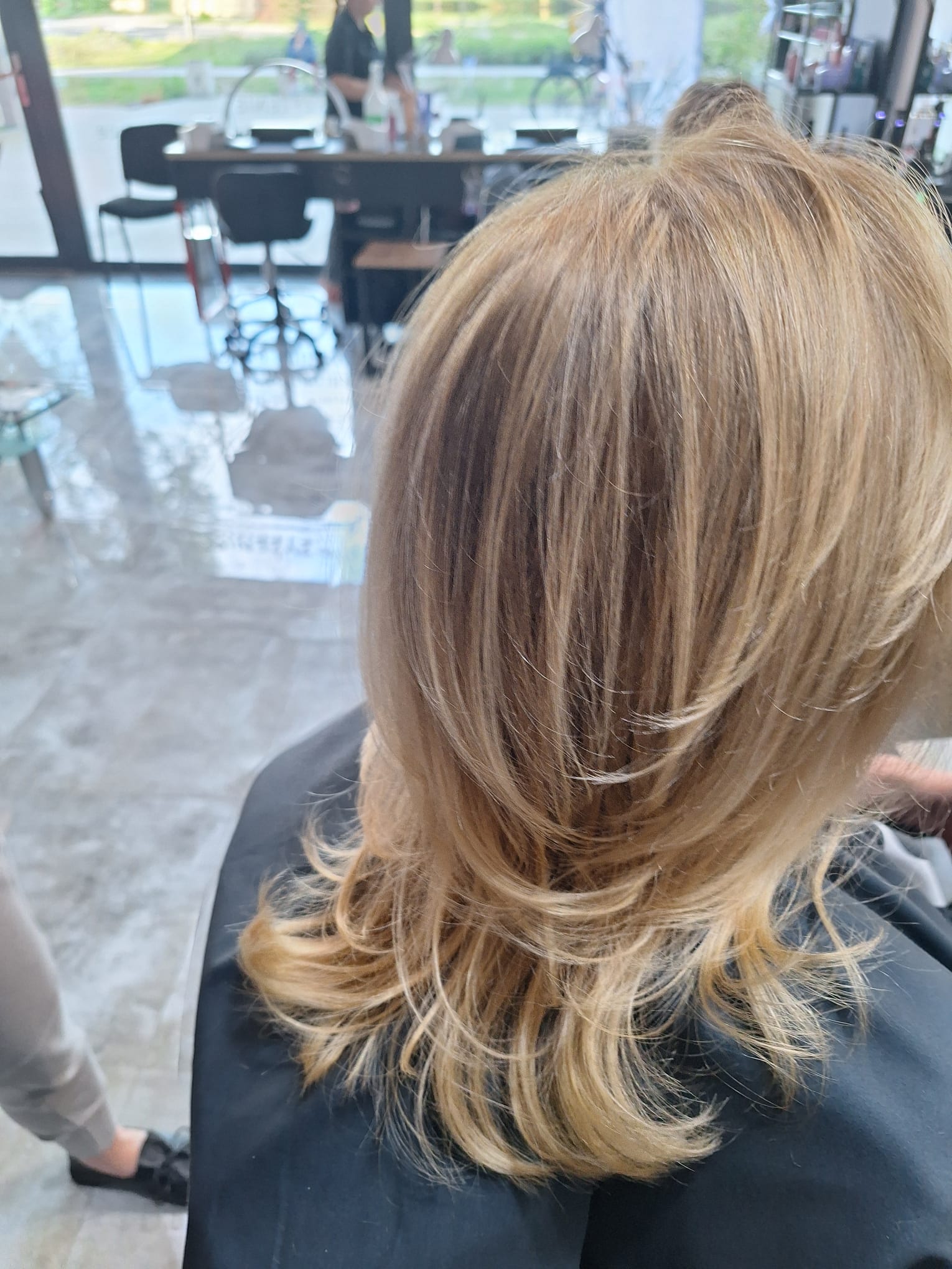 Warstwowe blond włosy z ciemniejszym odrostem i lekkim wywinięciem końcówek po stylizacji w salonie fryzjerskim.