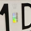 Zbliżenie na hologram zabezpieczający i znak orła umieszczone na białej polskiej tablicy rejestracyjnej pomiędzy czarnymi tłoczonymi znakami.